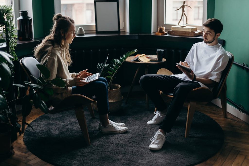 Two people sitting indoors discussing work, one using a laptop, the other with a tablet, in a modern, plant-filled room.