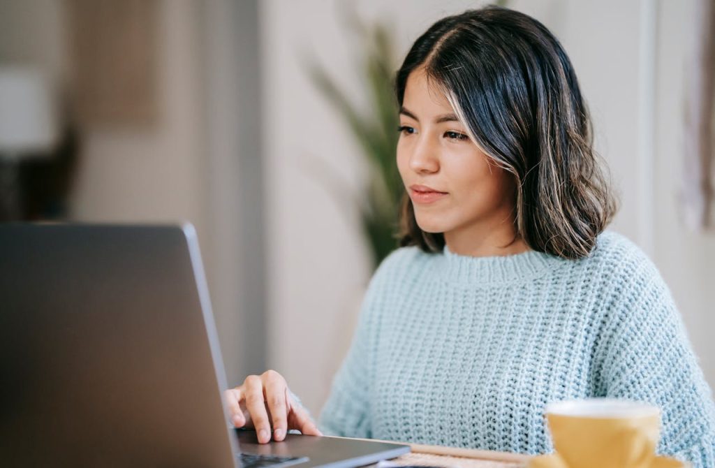 A confident young Latin American woman in casual clothes working remotely on a netbook at a table near a mug with coffee in a light apartment with a plant with green leaves near a wall